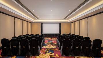 A conference room with rows of chairs, a carpeted floor, a projector screen, and a table with water bottles.