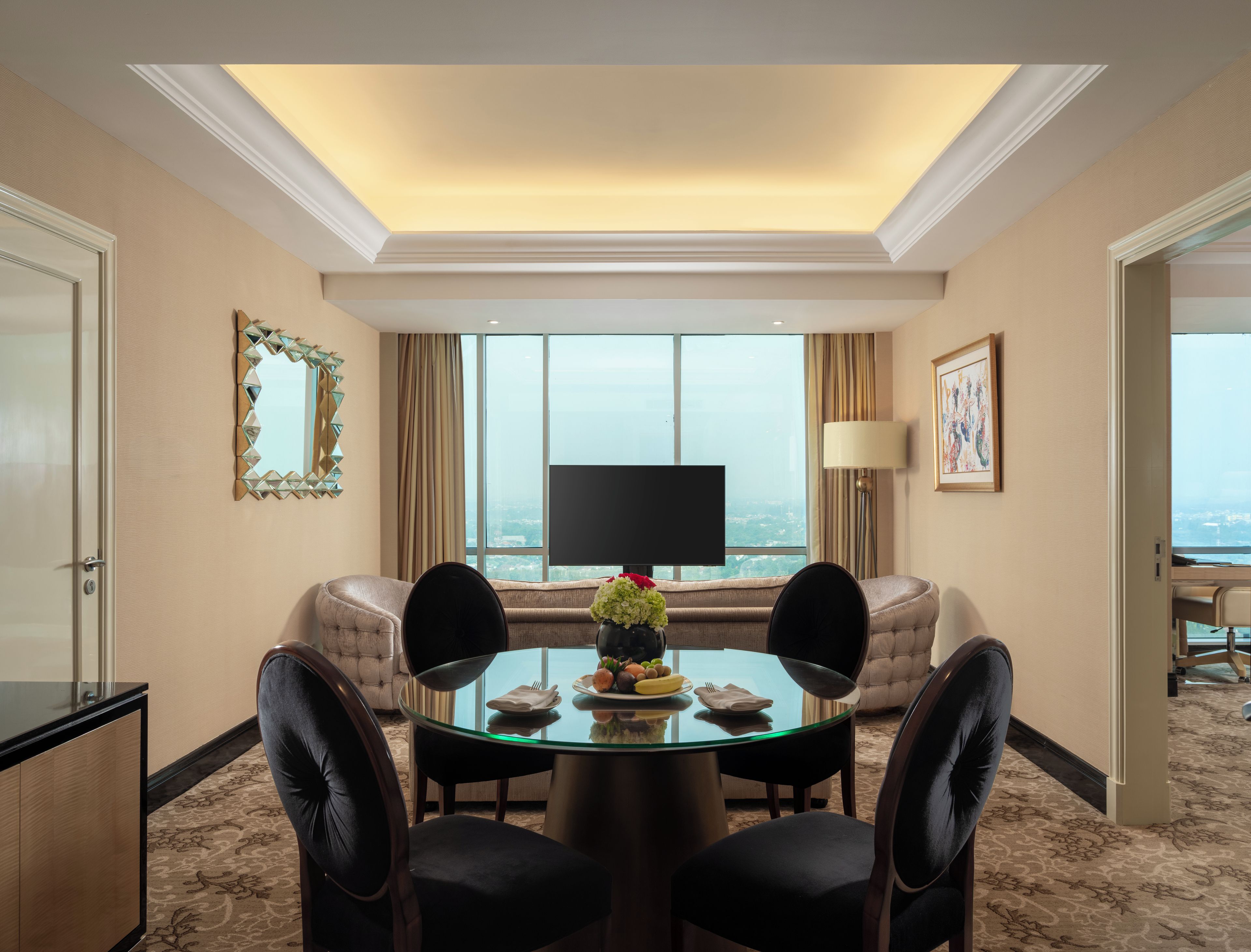 A hotel room with a dining area, a sofa, a TV, and a view of the city.