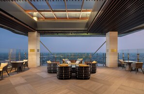 Un salón en la azotea con vistas panorámicas de la ciudad, que incluye una barandilla de cristal, muebles de mimbre y un diseño de techo moderno.