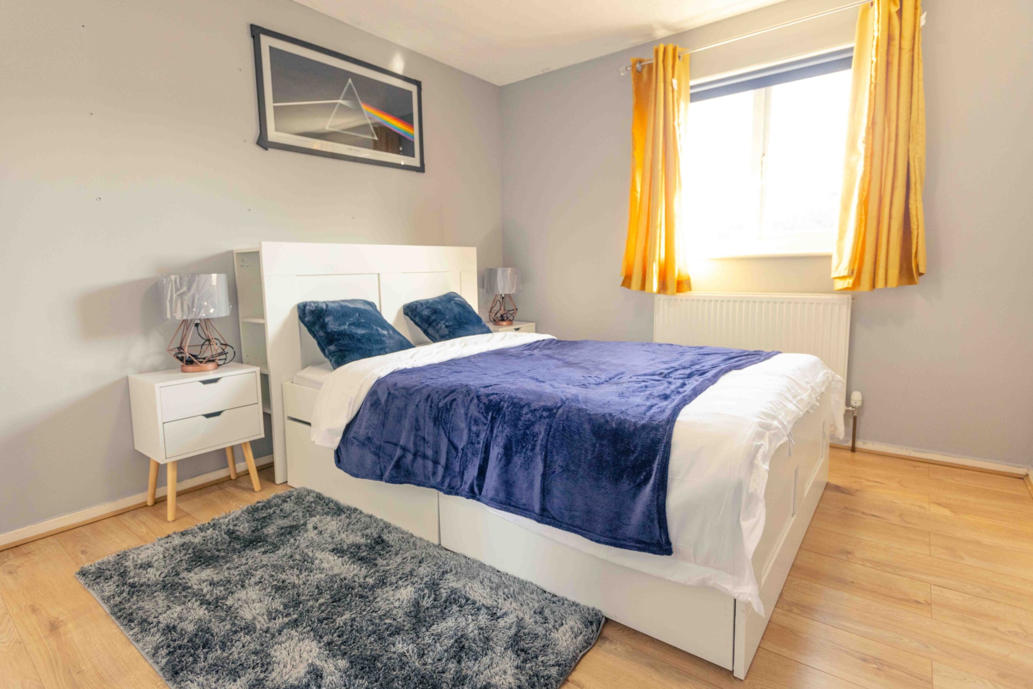 Un dormitorio con cama, mesitas de noche, una ventana con cortinas y un cuadro colgado en la pared.