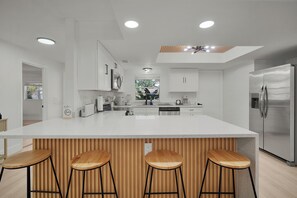 A modern kitchen with a large island, wooden bar stools, stainless steel appliances, and recessed ceiling lighting.
