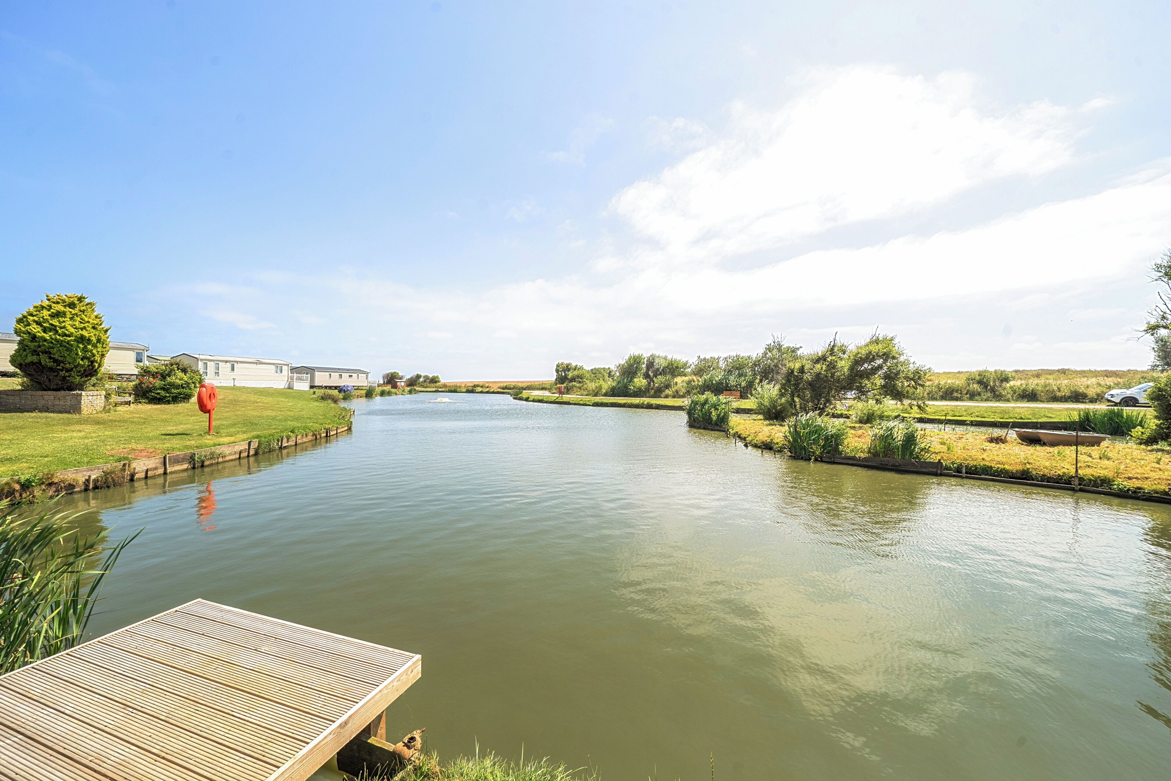 A calm river with a wooden dock, grassy banks, and a row of houses in the background.