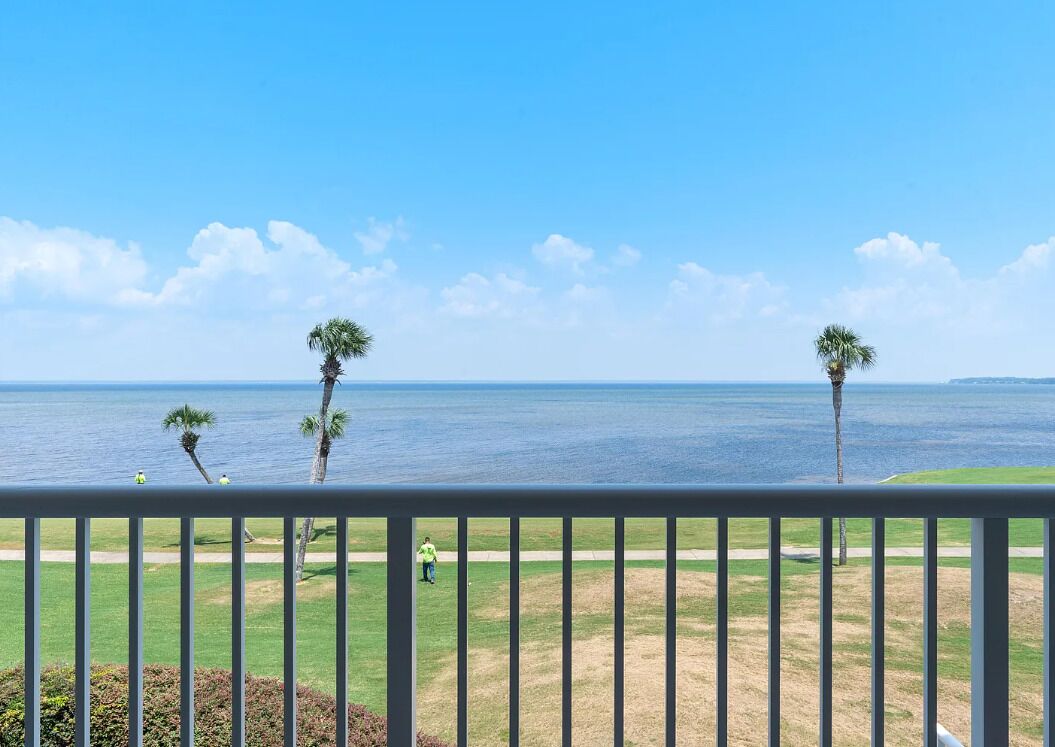 A balcony with a view of the ocean, palm trees, and a grassy area.