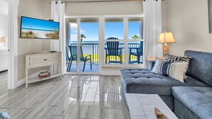Een moderne woonkamer met een grijze bank, een flatscreen televisie en een balkon met blauwe stoelen.