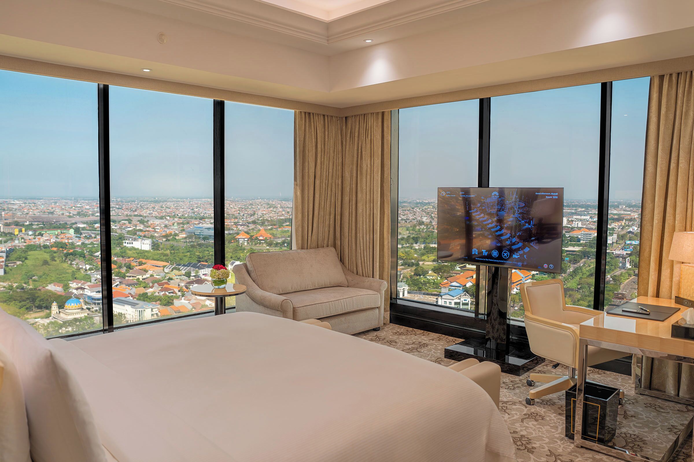 A hotel room with a large bed, a desk, a chair, and a TV. The room has large windows with a city view.