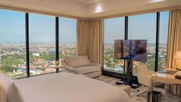 A hotel room with a large bed, a desk, a chair, and a TV. The room has large windows with a city view.