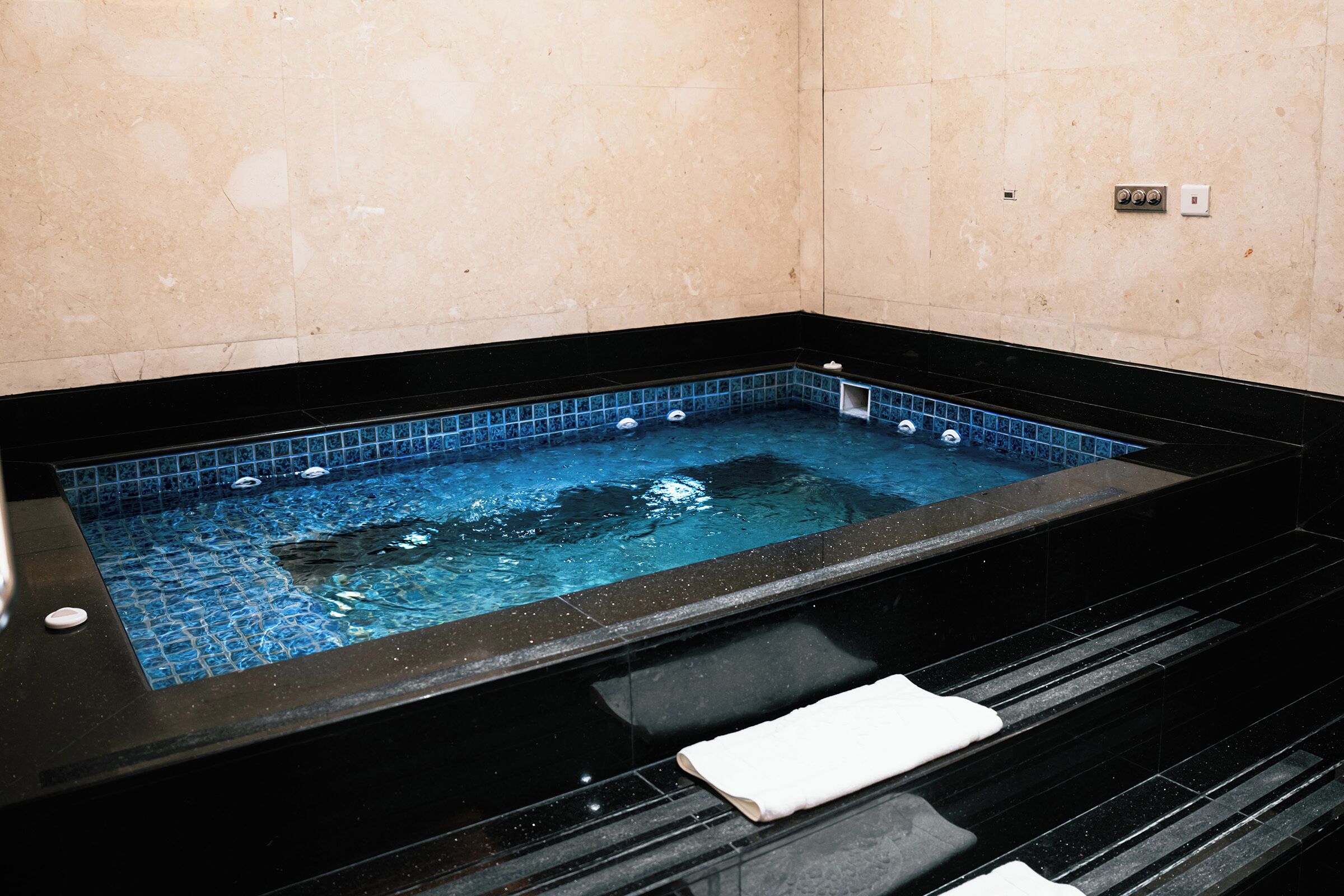 Un jacuzzi con borde negro y azulejos azules, una toalla blanca y un panel de control en la pared.