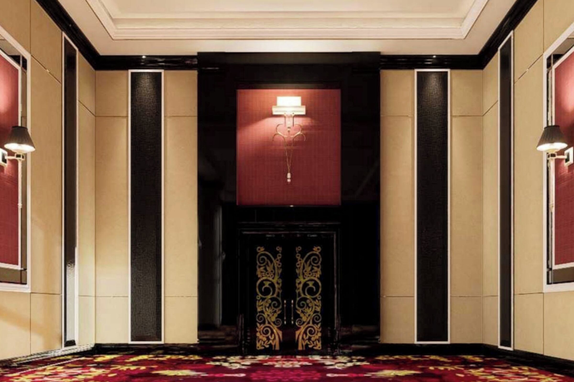 A hotel lobby with a patterned carpet, a dark door with ornate gold detailing, and wall-mounted lamps.