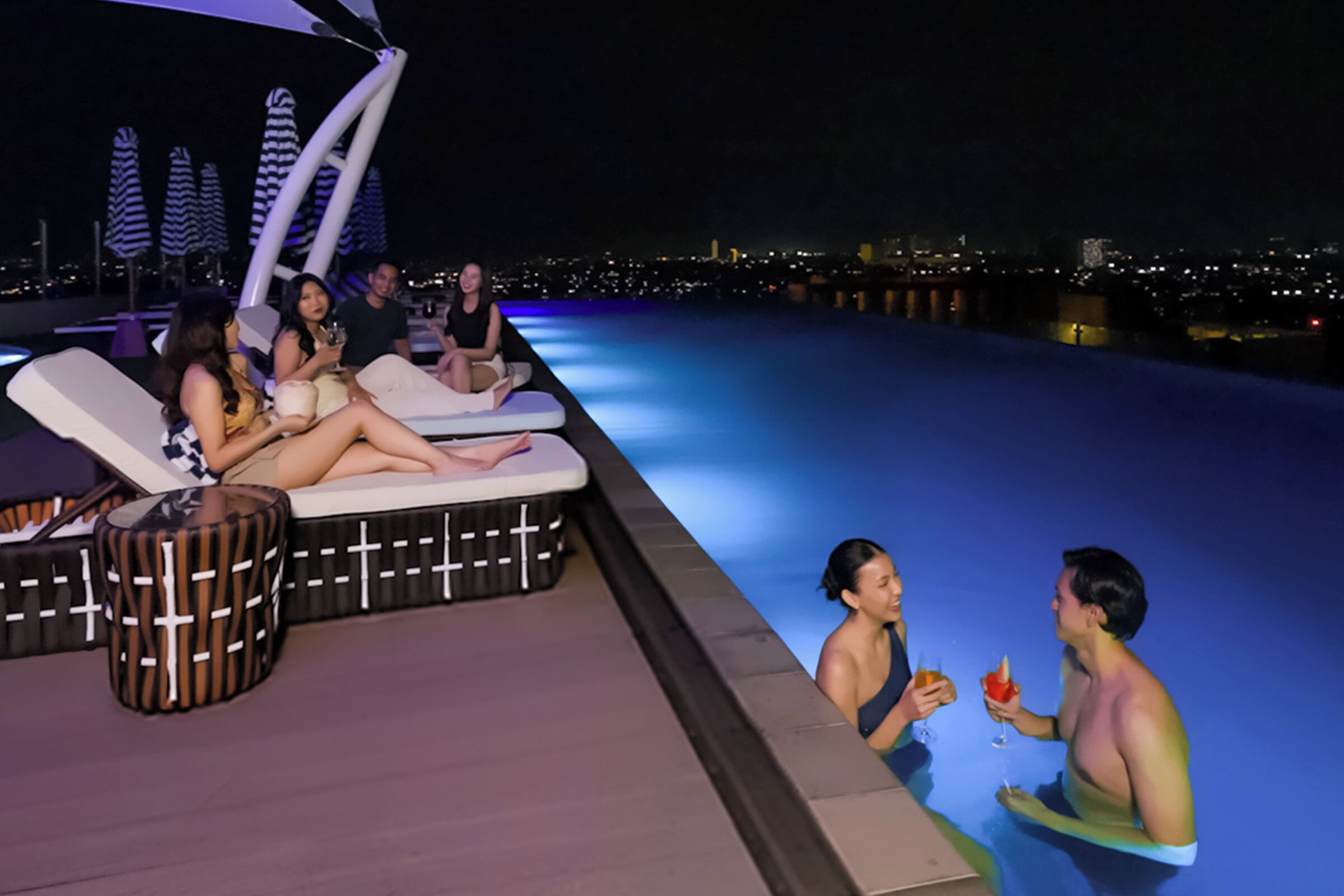 A rooftop pool area with lounge chairs and a cityscape view at night.