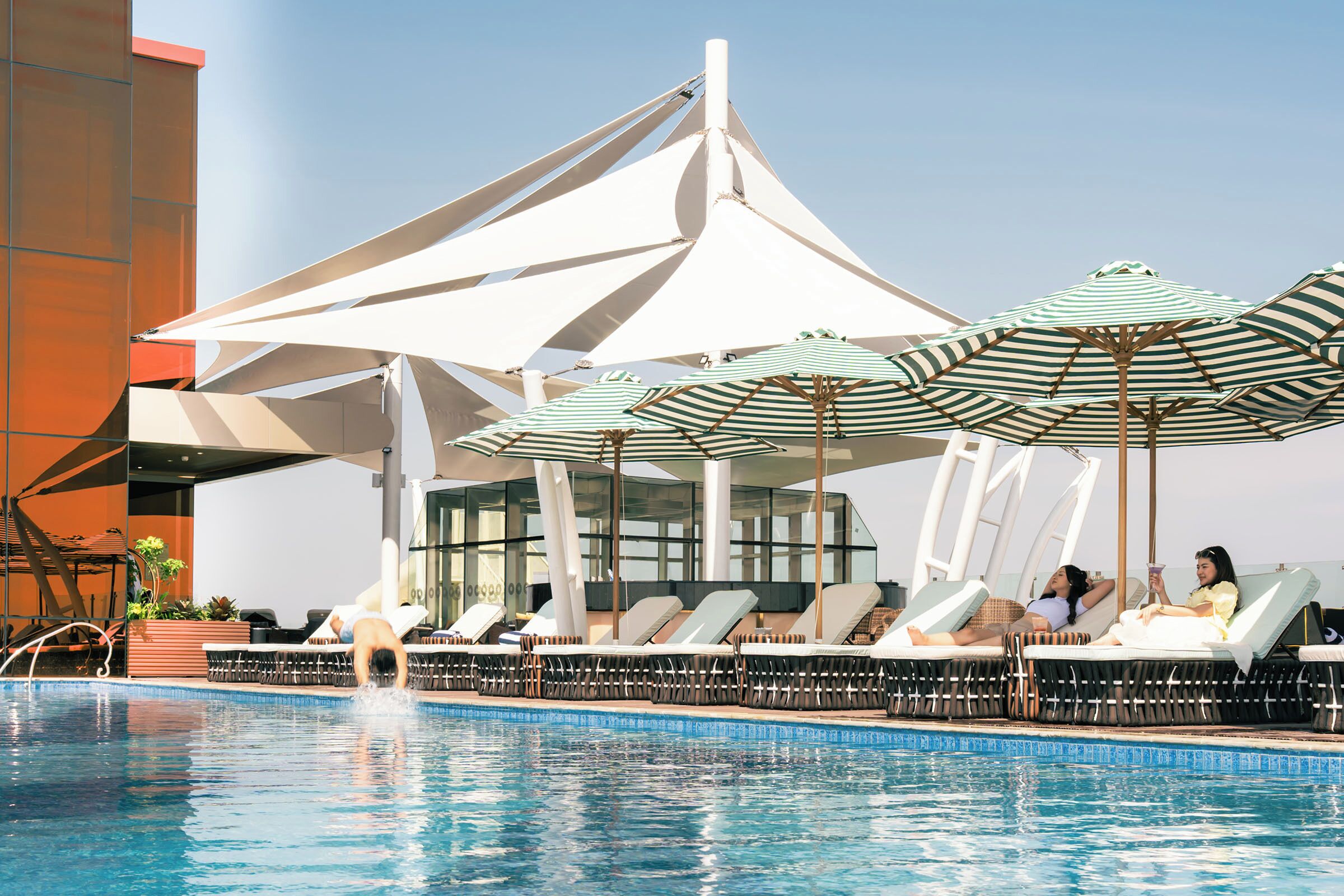 Uma área à beira da piscina com espreguiçadeiras e guarda-sóis, céu claro e uma estrutura de prédio moderna.