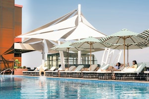 Zona de piscina con sillones de descanso y sombrillas, cielo despejado y una estructura de edificio moderna.