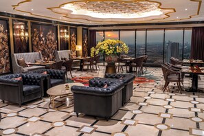 Un lobby de hotel lujoso con techo alto, decoraciones de pared elaboradas, un amplio ventanal con vista a la ciudad y una variedad de opciones de asiento, incluyendo sofás y sillones.