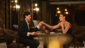 A couple dining at a table with a candle and a glass of red wine, in a room with wooden decor and ambient lighting.