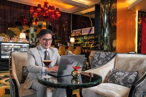 Un homme est assis à une table ronde, devant un ordinateur portable, avec un verre de martini et un vase décoratif. La pièce est décorée avec goût, avec un lustre, des fauteuils confortables et un coin bar à l’arrière-plan.