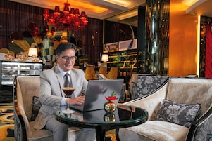 Un hombre sentado a una mesa redonda con una computadora portátil, una copa de martini y un jarrón decorativo. La habitación tiene una decoración elegante con una araña, asientos lujosos y un área de bar al fondo.