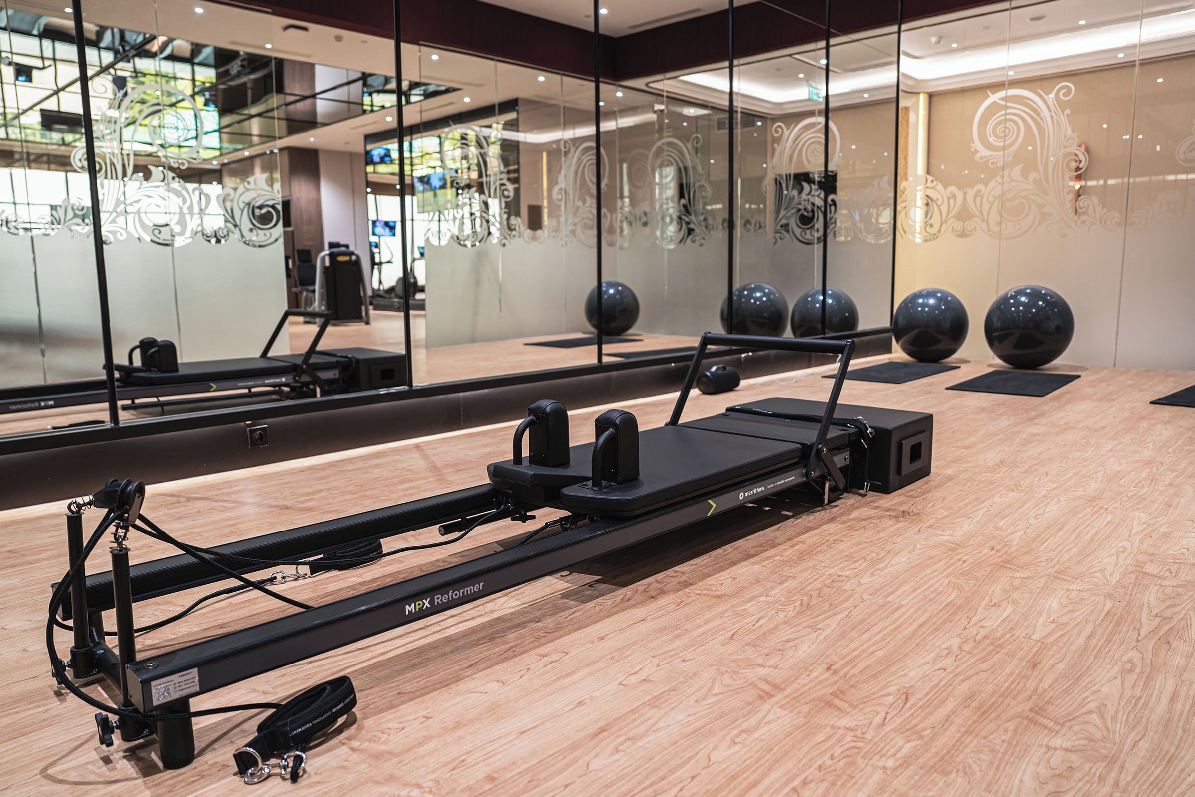 A Pilates reformer machine in a room with a wooden floor, large mirrors, and exercise balls.