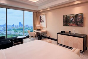 A hotel room with a large bed, a desk, a chair, a small table, and a view of the city.