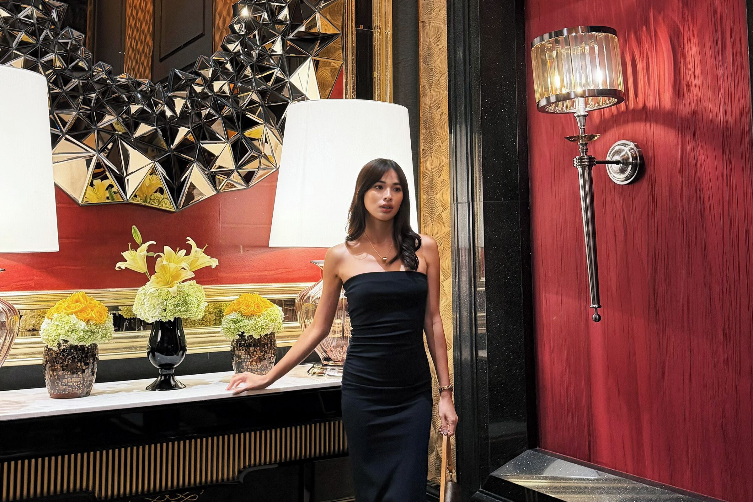 A woman standing in a room with a red wall, a large mirror, and a table with floral arrangements.