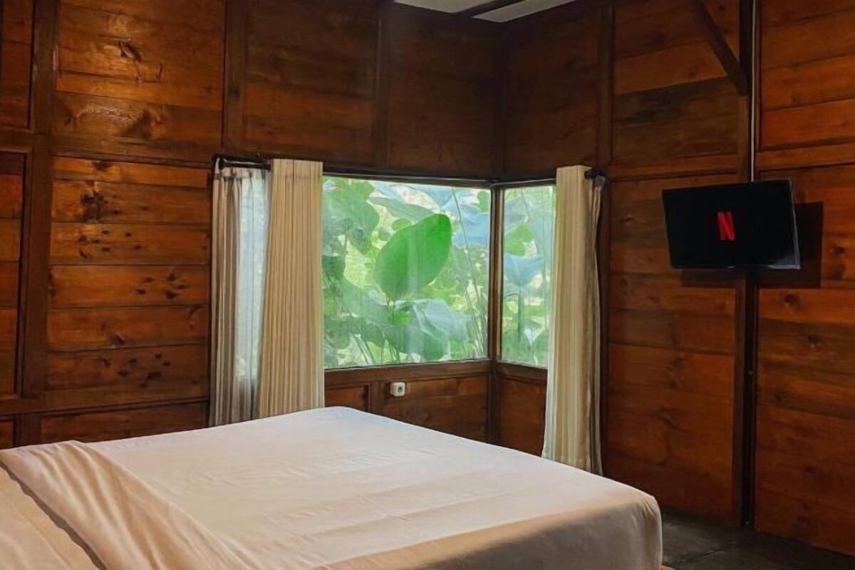 A wooden cabin room with a bed, a window with curtains, and a TV mounted on the wall.