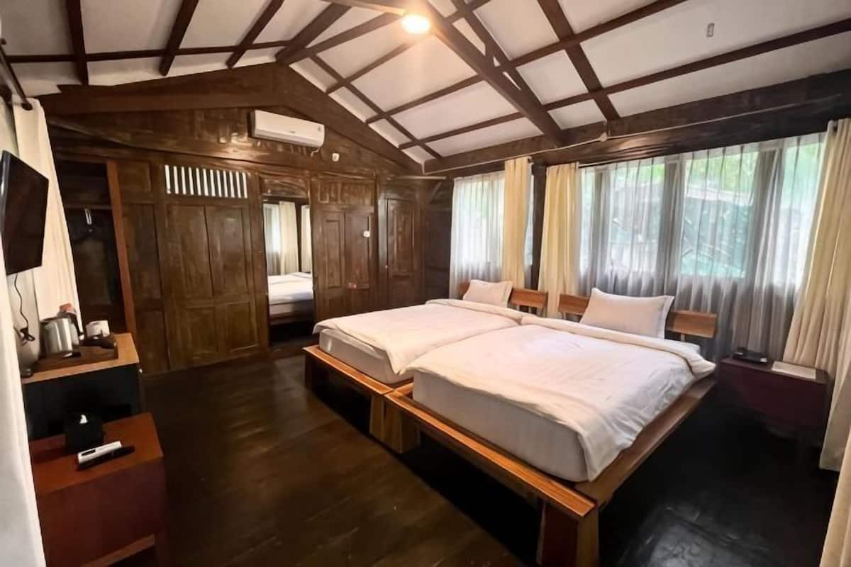 A room with a wooden ceiling, two beds, a TV, and a window with curtains.