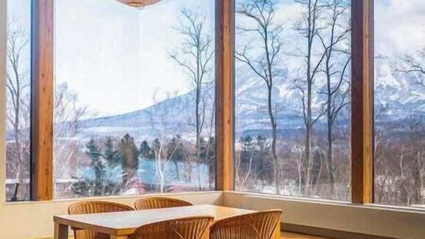A dining area with a table and chairs, large windows with a view of a mountainous landscape. - Aoyama Lodge | 4BR Chalet in Kabayama | Stunning Mt. Yotei Views (Kutchan-cho)