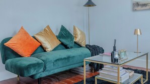 A teal sofa with multicolored cushions, a glass coffee table with books, a black floor lamp, and a framed picture on the wall. - Rooftop/worktable/Netflix/Parking/Promo/longstay (London)