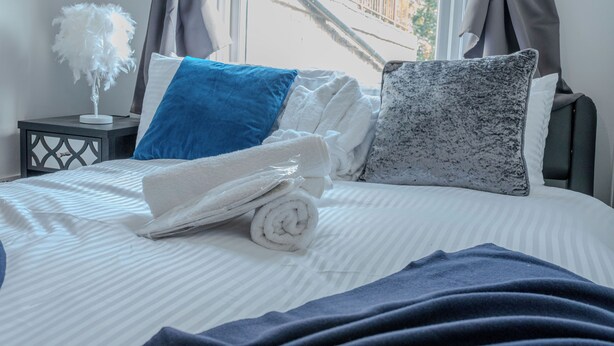 A neatly made bed with rolled towels, pillows, and a decorative feathered object on a bedside table. - Rooftop/worktable/Netflix/Parking/Promo/longstay (London)