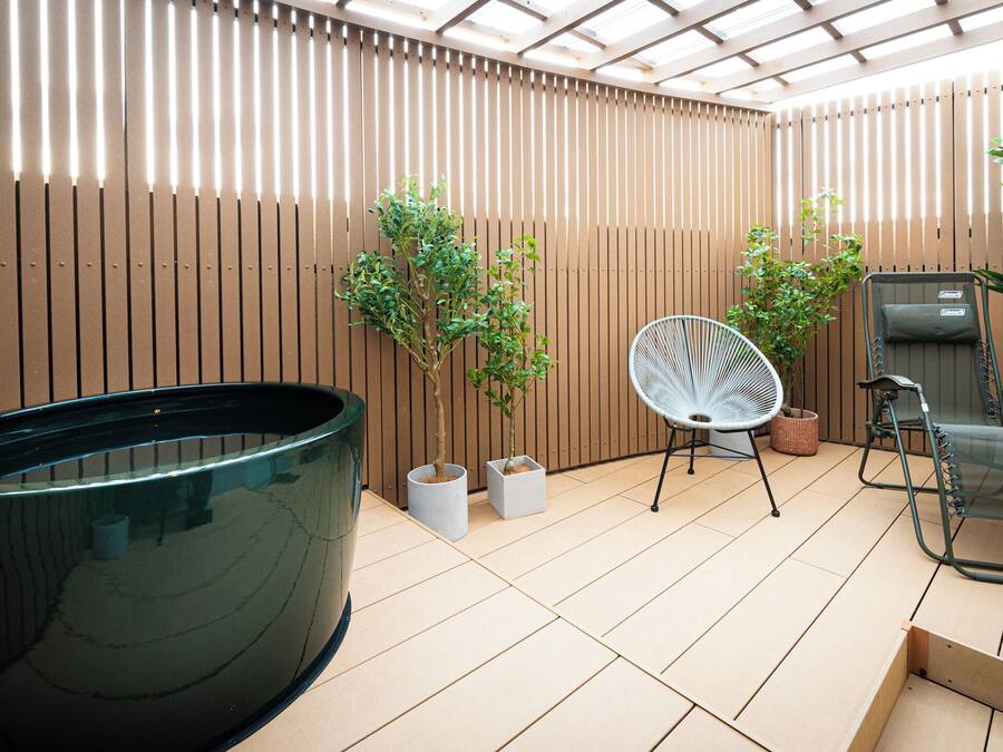 A modern outdoor bathroom with a large black bathtub, two potted plants, a chair, and a small table.