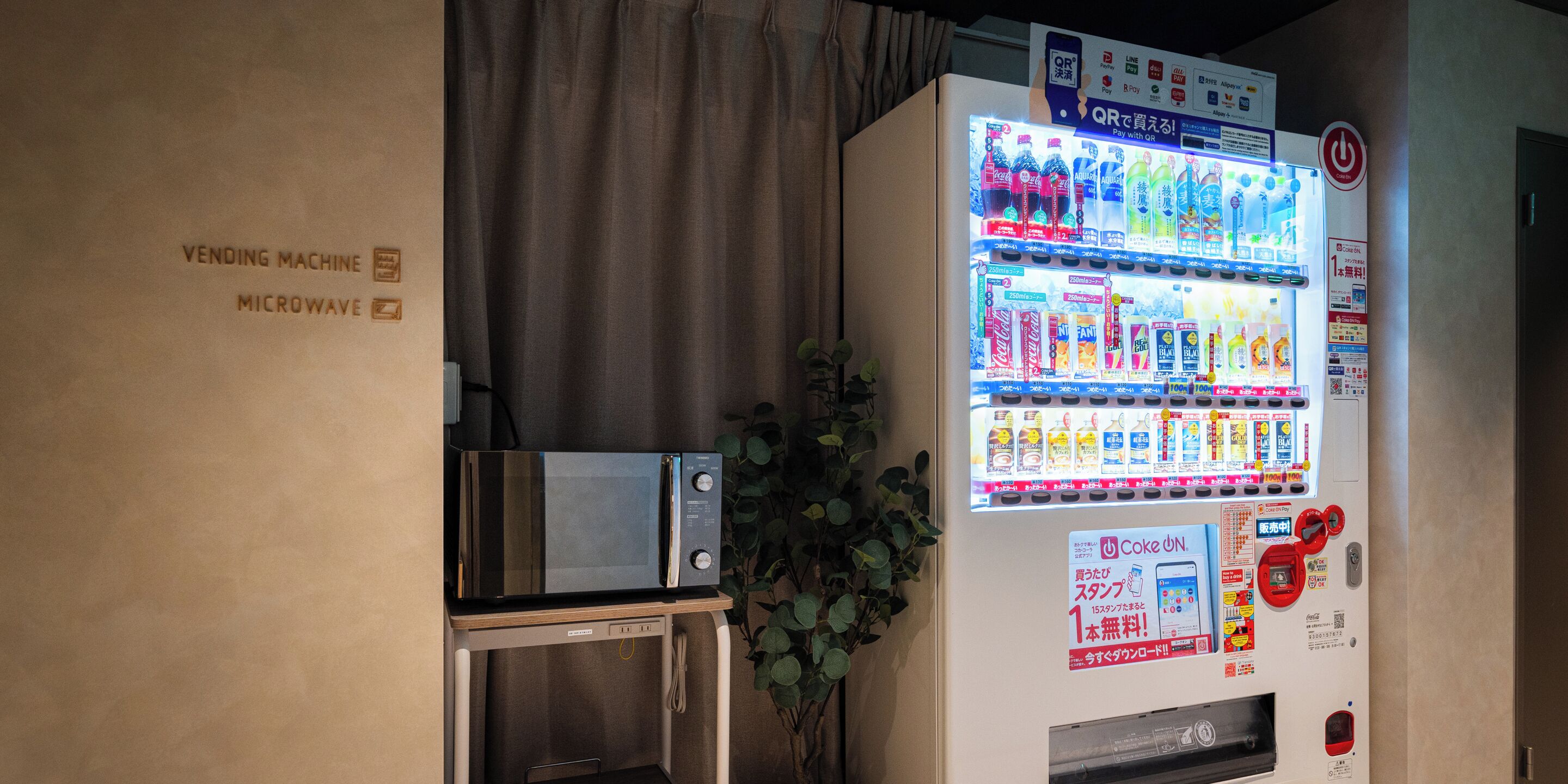 Ein Getränkeautomat mit verschiedenen Getränken und Snacks, eine Mikrowelle und ein Schild mit einem QR-Code für die Bezahlung.