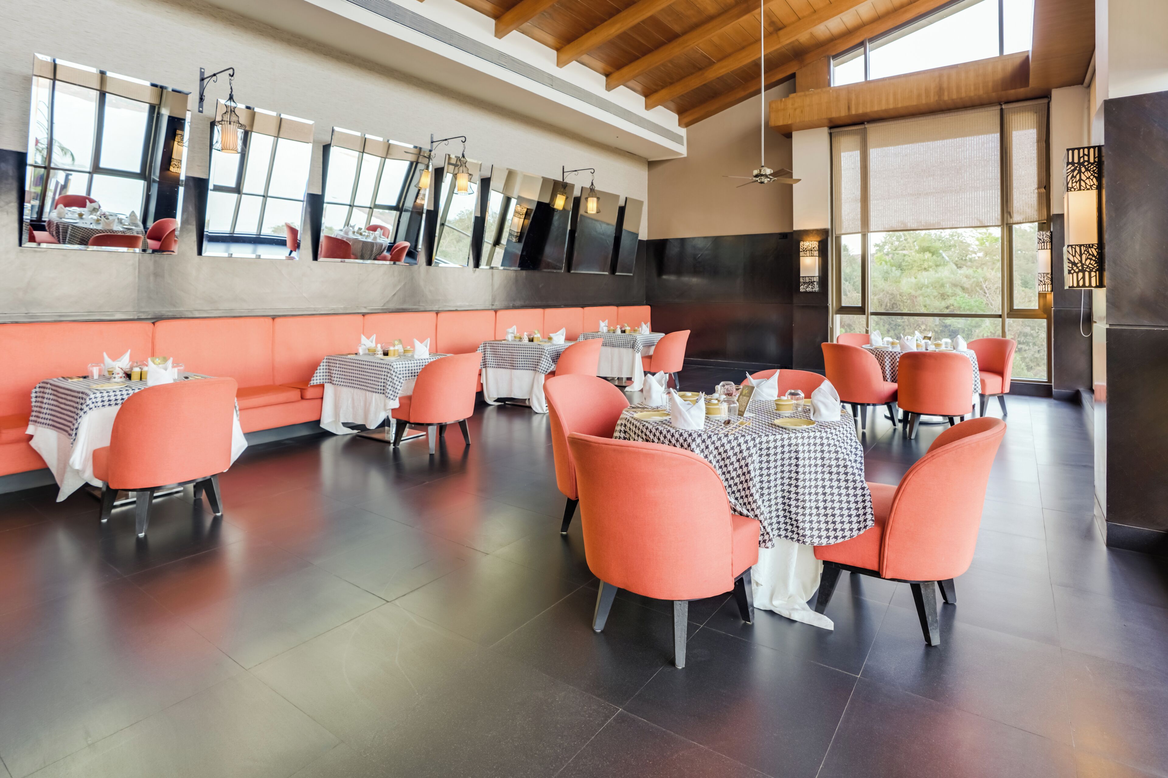 A spacious restaurant with high ceilings, large windows, and orange seating. Tables are set with white tablecloths and silverware.