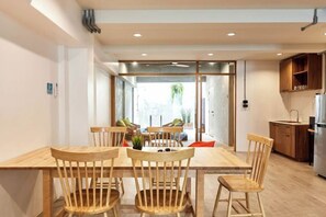 A modern kitchen with wooden cabinets, a dining area with a wooden table and chairs, and a view through a glass door to an outdoor area. - Vann House Hua Hin (Hua Hin)