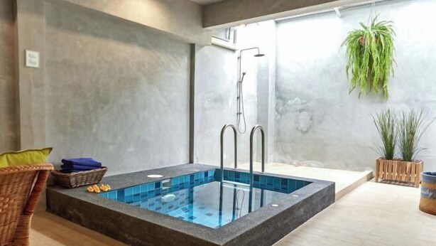 Un área de piscina interior moderna con piscina revestida de vidrio, una ducha y una silla de mimbre con toallas.