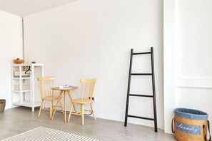 Une pièce avec un mur blanc, une échelle noire, deux chaises en bois, une petite table ronde, une étagère blanche et un panier à linge bleu.
