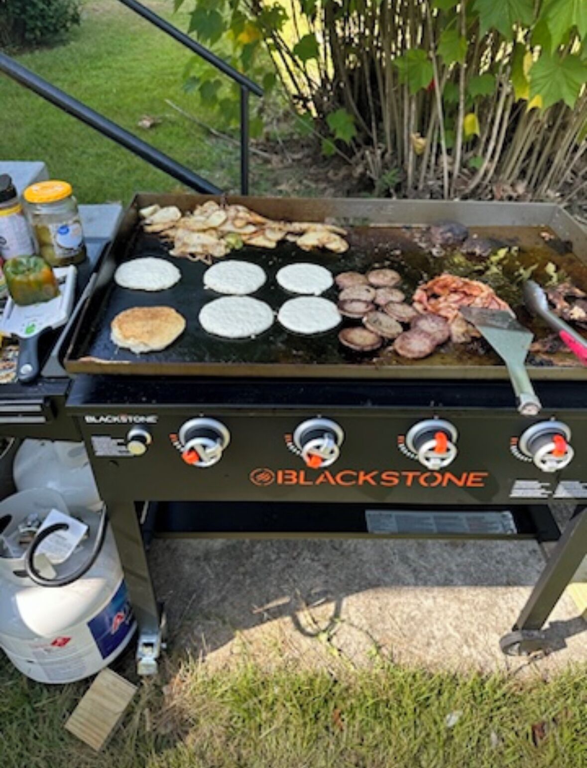 A Blackstone grill with multiple cooking surfaces, including patties and sausages, set outdoors.
