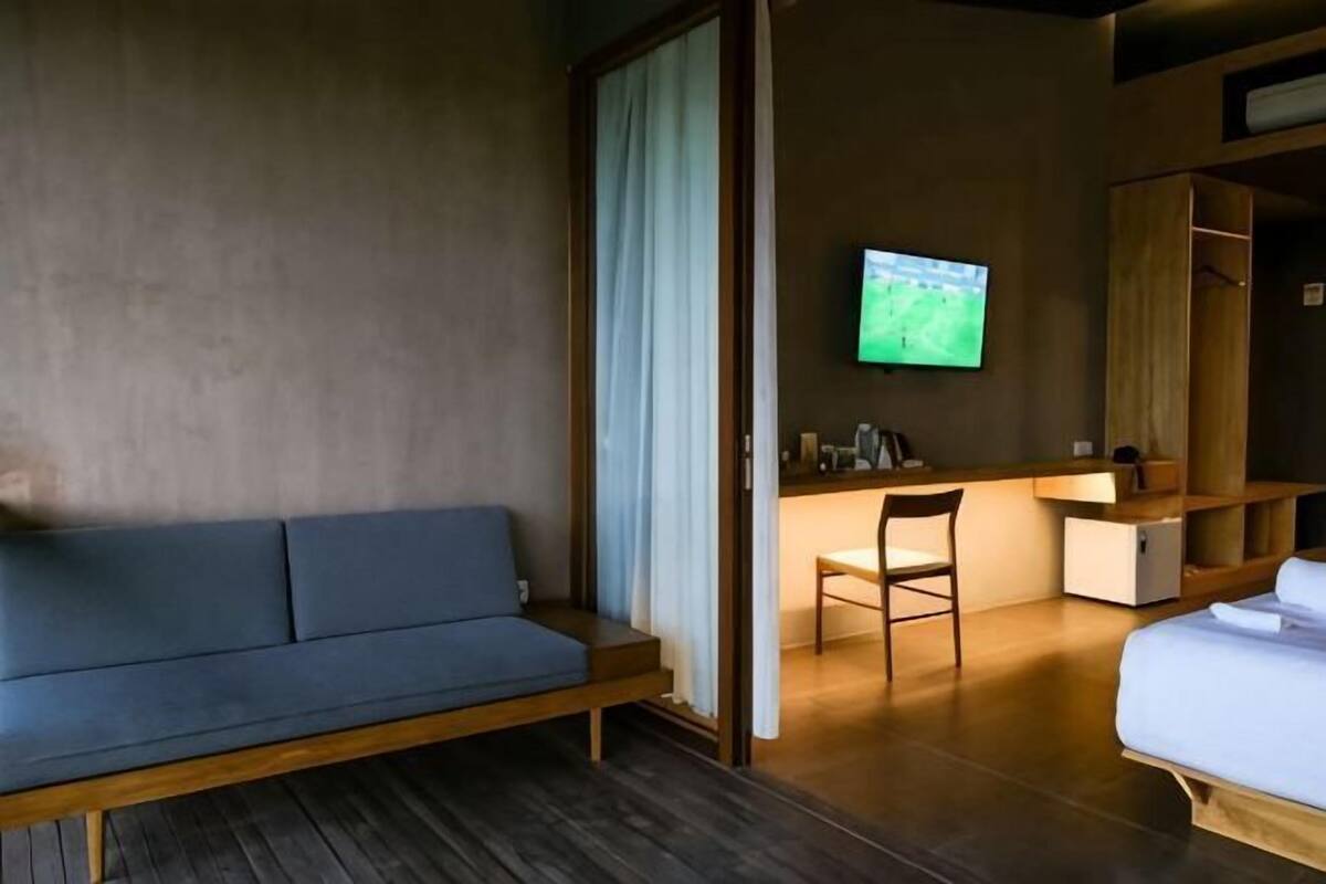 A room with a wooden floor, a blue sofa, a desk with a chair, a TV mounted on the wall, and a bed.