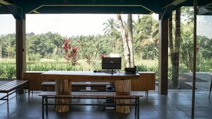 Un espace de restauration extérieur abrité, avec une table et des bancs en bois, offrant une vue sur un paysage luxuriant, avec des palmiers et une rizière.