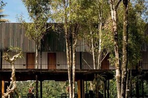 Una casa en el árbol con terraza de madera y barandilla metálica, rodeada de árboles.