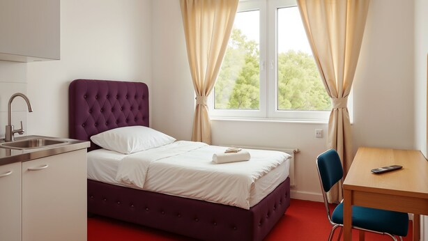 A bedroom with a purple bed, white bedding, a wooden desk, a chair, a window with curtains, and a sink. - King Hotel (Huddinge)