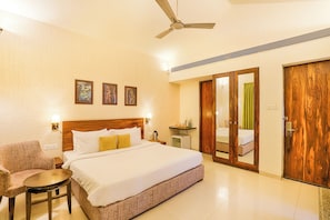 A hotel room with a bed, a chair, a small table, a ceiling fan, and a wooden wardrobe. - Tropicana Resort & Spa Alibaug (Alibag)