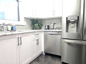 A modern kitchen with white cabinets, a stainless steel refrigerator, a dishwasher, and a window providing natural light. - Modern 2BR Haven - Walk to Transit, Dining & Entertainment (Edmonton)