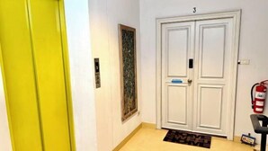 A hallway with a yellow door, a white double door with the number 3, a framed artwork on the wall, a fire extinguisher, and a black chair. - Samsen Suites (Bangkok)