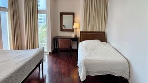 A hotel room with two single beds, a desk, a chair, a lamp, and a window with curtains. - Samsen Suites (Bangkok)