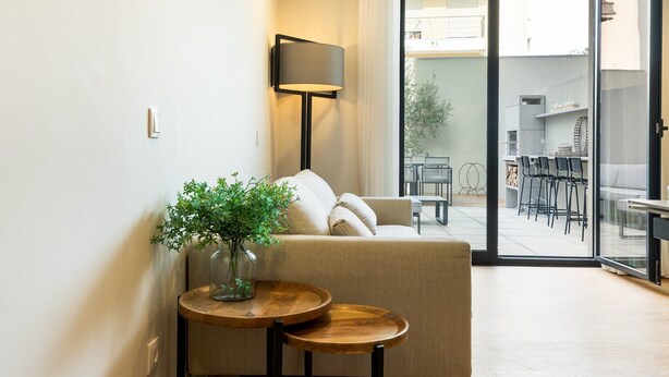 Ein modernes Wohnzimmer mit einer Couch, einem kleinen Holztisch mit einer Vase mit Grünpflanzen und einer Stehlampe. Eine Glastür ermöglicht den Zugang zu einem Außenbereich mit einem Esstisch und Stühlen.