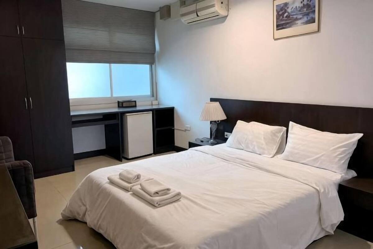 A hotel room with a bed, a desk, a chair, a nightstand, and a window with blinds.