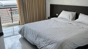 A hotel room with a large bed, a nightstand, and a view of a building through a window with curtains. - Tongyang House Serviced Apartment (Bangkok)