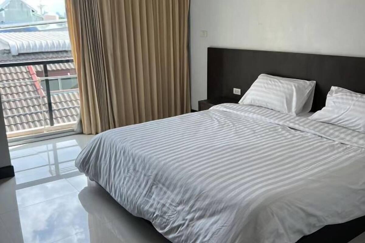 A hotel room with a large bed, a nightstand, and a view of a building through a window with curtains.