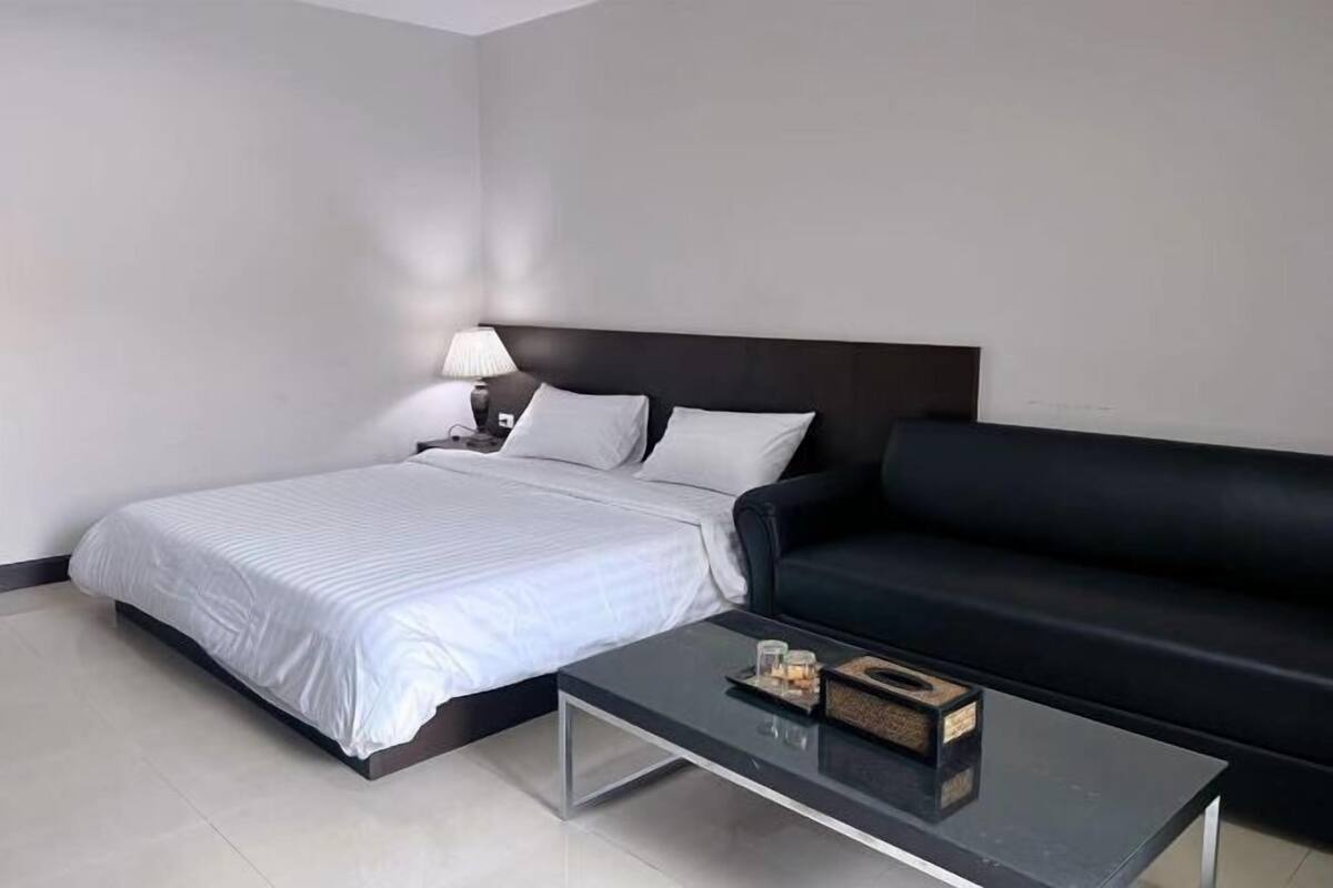 A modern hotel room with a bed, a black sofa, a glass coffee table, and a nightstand with a lamp.