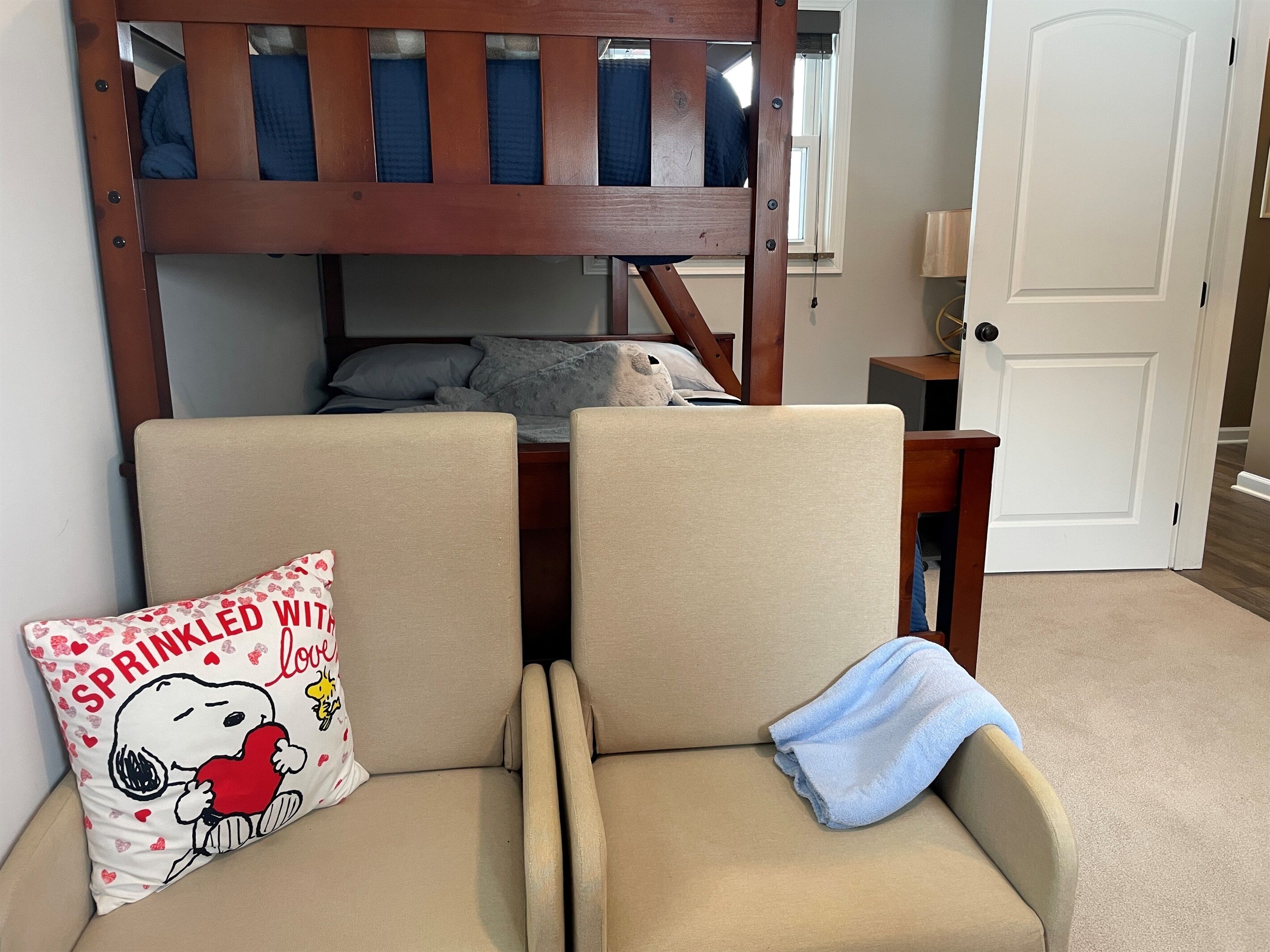 Een stapelbed met hoofdeinde, twee beige fauteuils, een kussen met Snoopy-print en een blauwe handdoek.