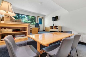 A room with a wooden table, four chairs, a bookshelf, a TV mounted on the wall, and a window with a view of trees.