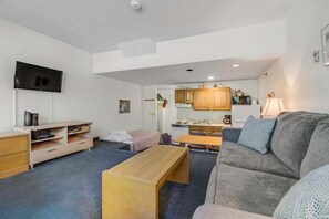 A compact living space with a sofa, a wooden coffee table, a TV mounted on the wall, and a kitchenette in the background.